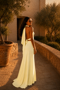 Woman in a yellow dress standing in a sunlit outdoor setting with trees and a building.